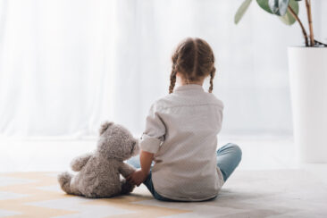 Una niña pequeña está sentada en el suelo, vista desde atrás. Tiene en la mano un osito de peluche gris.