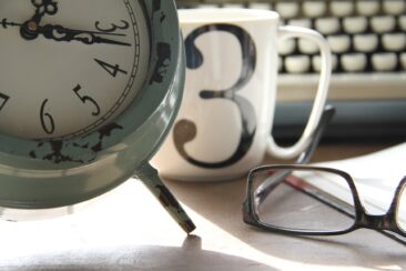 A clock, a cup with the number 3 and a pair of glasses are lying on a table in front of a typewriter.