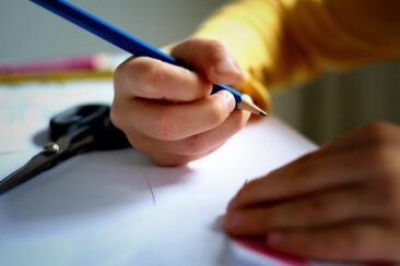 Children's hands hold a blue pencil over a sheet of paper.